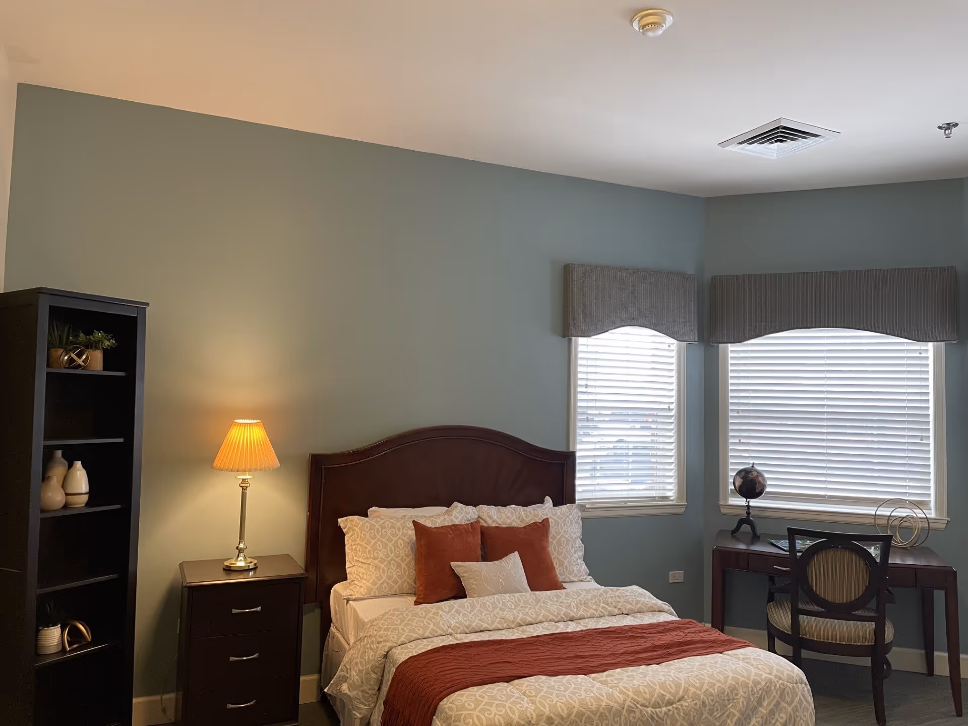 A cozy bedroom with a neatly made bed featuring white and rust-colored bedding and pillows. To the left of the bed is a dark wooden nightstand with a lamp on it, and next to that is a tall black bookshelf with decorative items. On the right side of the room, there is a wooden desk with a chair, a small globe, and decorative objects placed near two windows with blinds and valances. The walls are painted a soft green color.