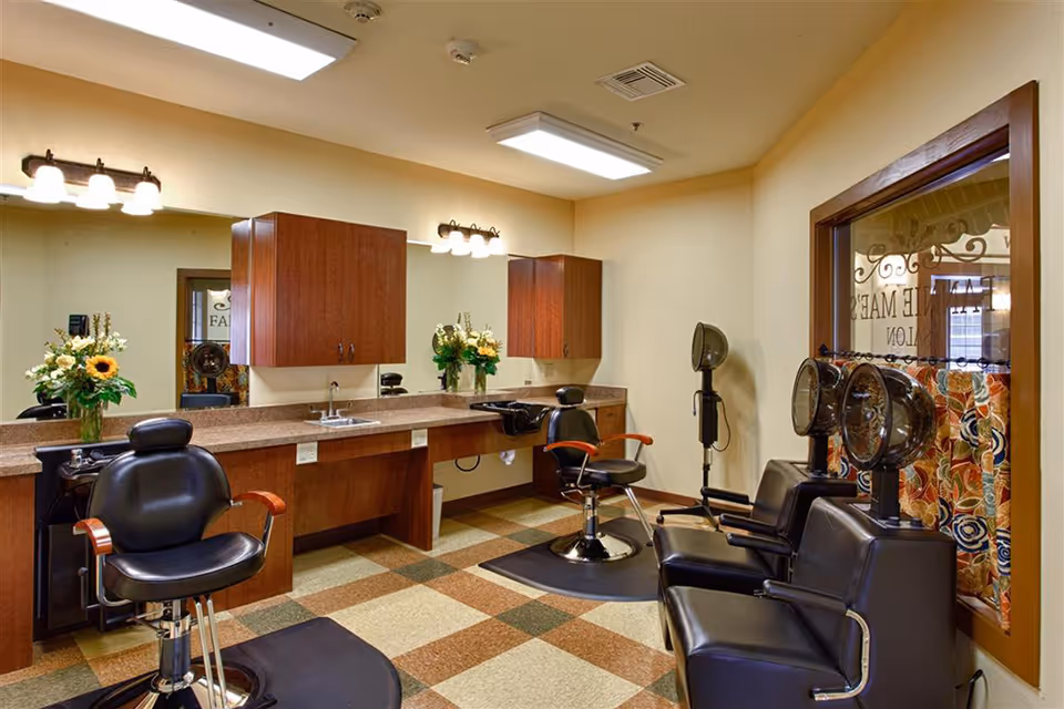 Interior view of a salon area with black salon chairs, hair dryers, a large mirror, wooden cabinets, and floral arrangements on the counter. The room has a checkered tile floor and warm lighting.