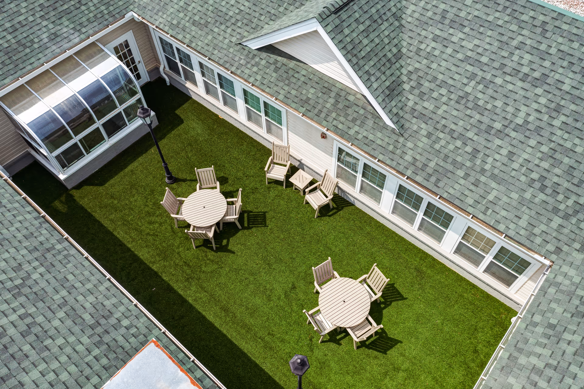 Aerial view of a courtyard with artificial grass, two round tables each surrounded by four wooden chairs, additional wooden chairs and small tables along the building walls, and two black lampposts. The courtyard is enclosed by a building with green shingled roofs and multiple windows.