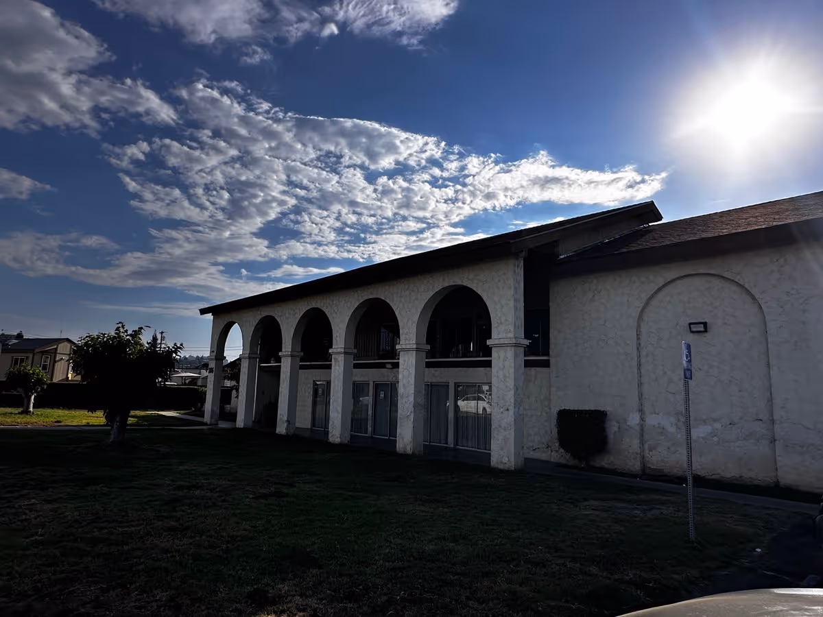Two-story stucco building with arched colonnade facing a grassy lawn under a bright sky.
