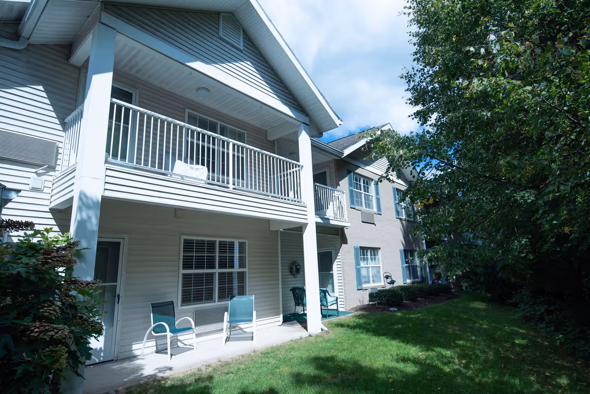 Two-story residential building with covered balconies, ground-level patios with chairs, and a grassy yard bordered by trees.