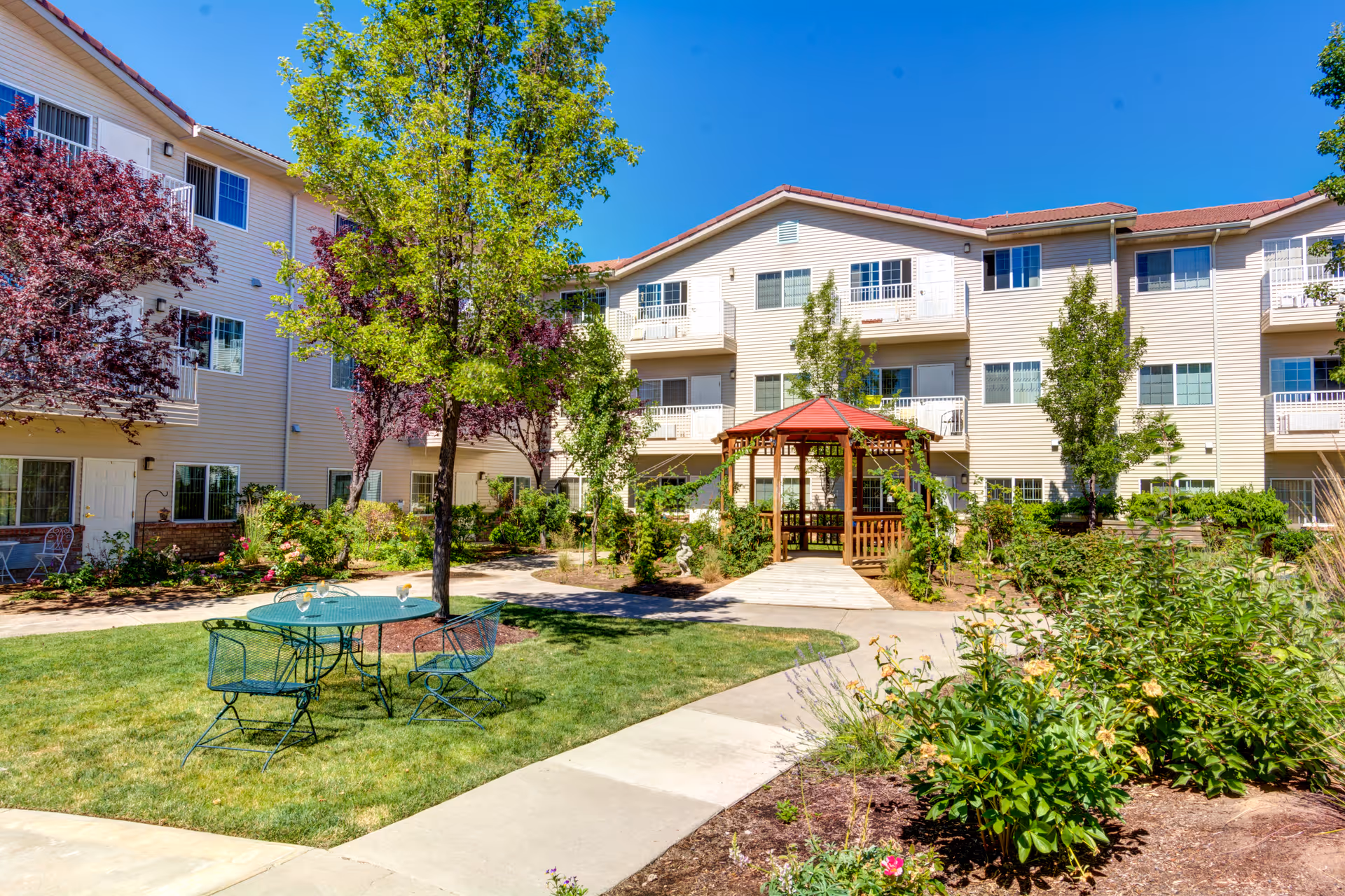 Outdoor courtyard area of a retirement community with a green metal table and chairs on a grassy patch, surrounded by trees and bushes. A wooden gazebo with a red roof is visible in the background, along with a three-story beige building with balconies and windows under a clear blue sky.
