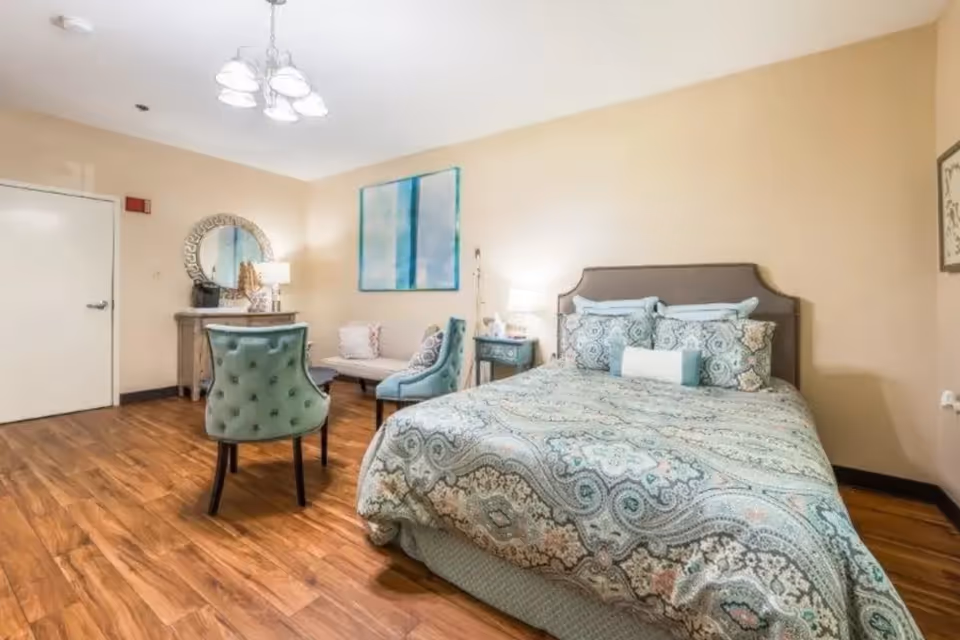 A cozy bedroom with a large bed covered in a patterned comforter and multiple pillows. The room features wooden flooring, a small sitting area with two blue upholstered chairs and a small couch, a wooden dresser with a round mirror above it, and soft lighting from table lamps and a ceiling light fixture.
