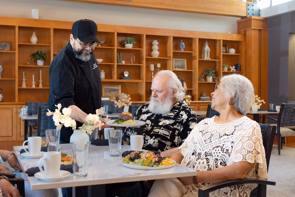 A server places a plate of food in front of two older adults seated at a dining table in a well-lit dining room with wooden shelving behind them.