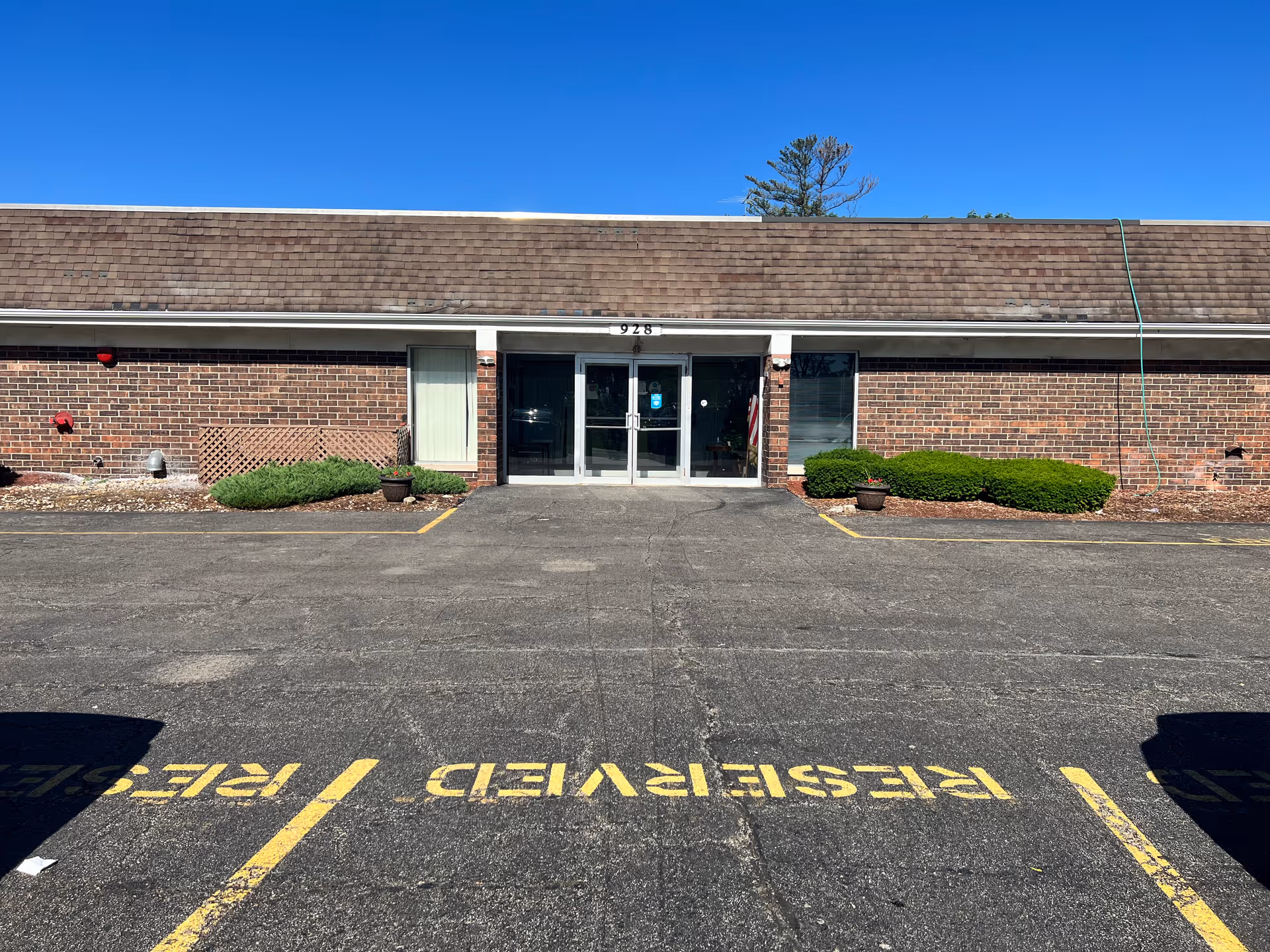 Exterior front view of a single-story brick building with a shingled roof under a clear blue sky. The building has a central glass double door entrance with the number 928 above it, flanked by windows and small bushes on either side. The foreground shows a paved parking lot with yellow painted lines and the word RESERVED marking parking spaces.