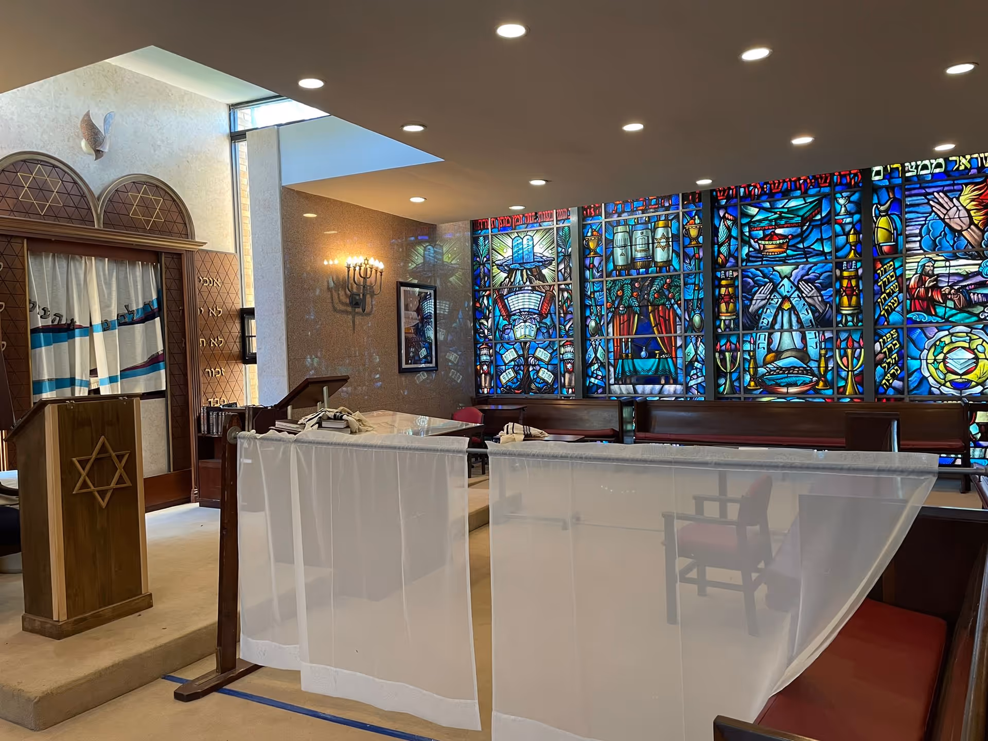Interior view of a synagogue or prayer room with a wooden podium featuring a Star of David, a curtain with Hebrew text, and a large stained glass window depicting religious symbols and scenes. The room has recessed ceiling lights and a wall-mounted menorah.