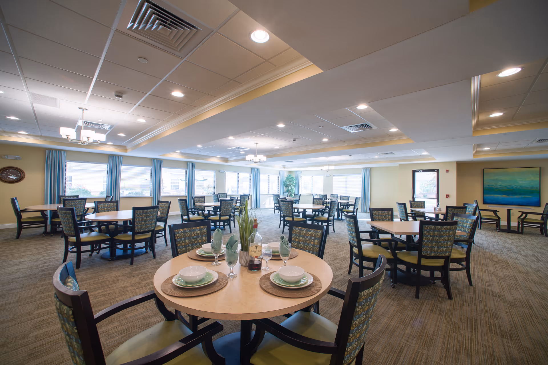 A spacious dining room with multiple round tables and chairs arranged neatly. One table in the foreground is set with plates, bowls, glasses, and napkins. The room has large windows with blue curtains allowing natural light to fill the space. The ceiling has recessed lighting and chandeliers, and there is a large painting on one wall.