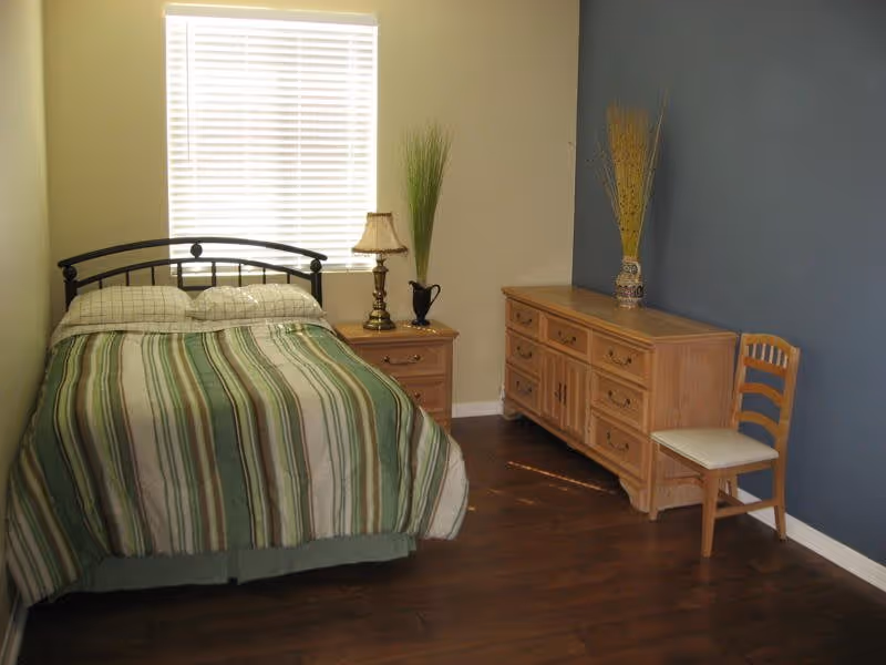 A bedroom with a bed covered in a green and beige striped comforter, a wooden nightstand with a lamp and a decorative vase, a wooden dresser with multiple drawers, a wooden chair with a white cushion, and a window with white blinds letting in natural light. The walls are painted beige and blue, and the floor is dark wood.