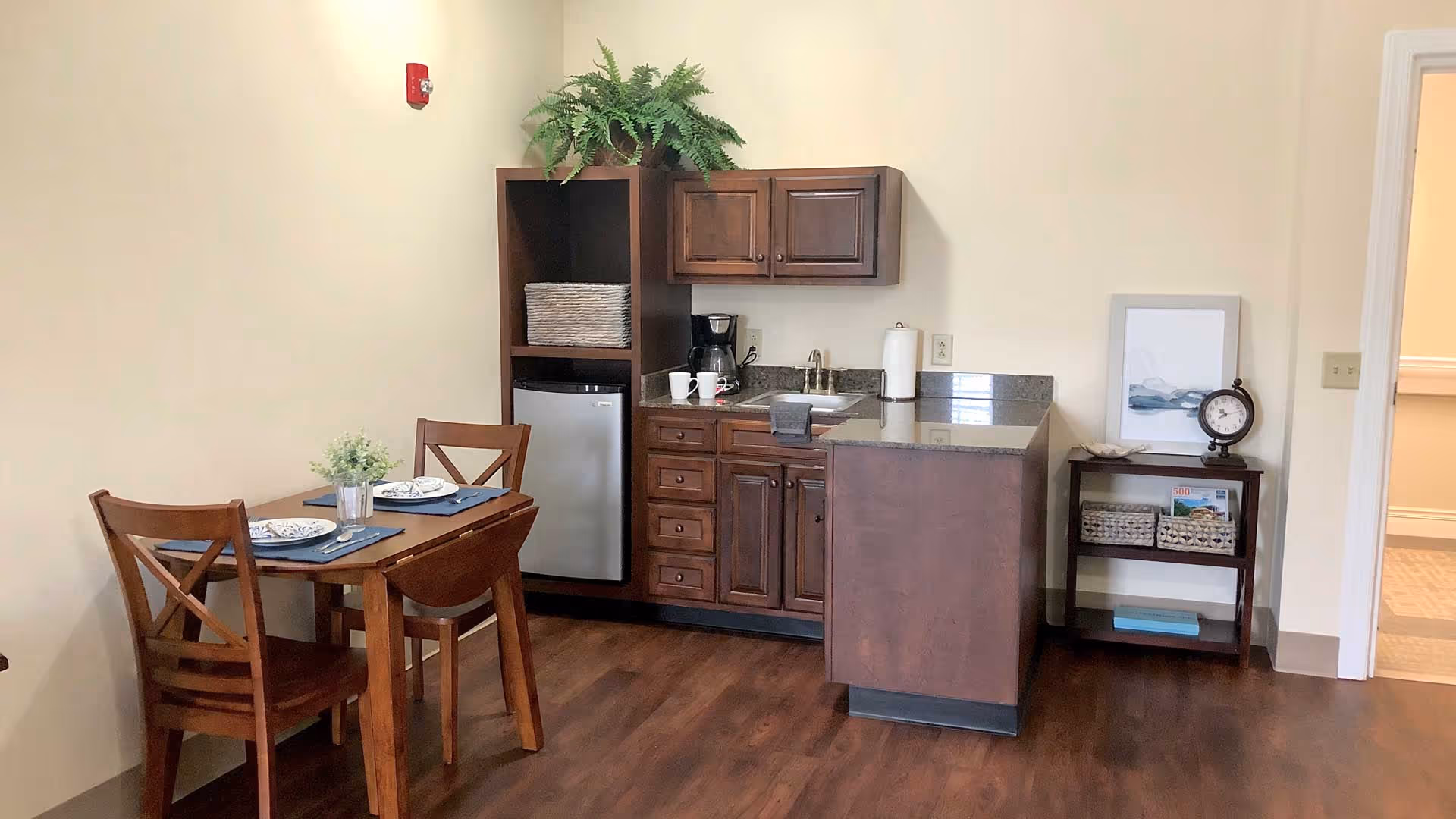 Small kitchenette and dining area with wooden cabinets, mini fridge, sink and a two-seat dining table in a furnished suite.