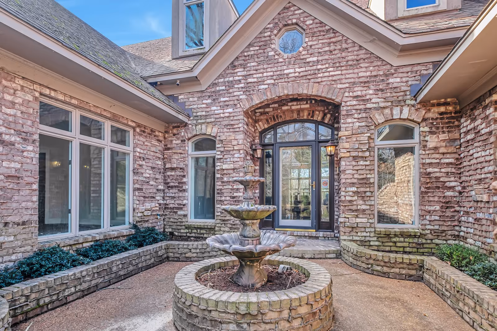 Outdoor courtyard area of a senior living facility with a three-tiered stone fountain in the center, surrounded by brick walls and windows. The entrance door with glass panels and two wall-mounted lantern lights is visible in the background under an arched brick design.