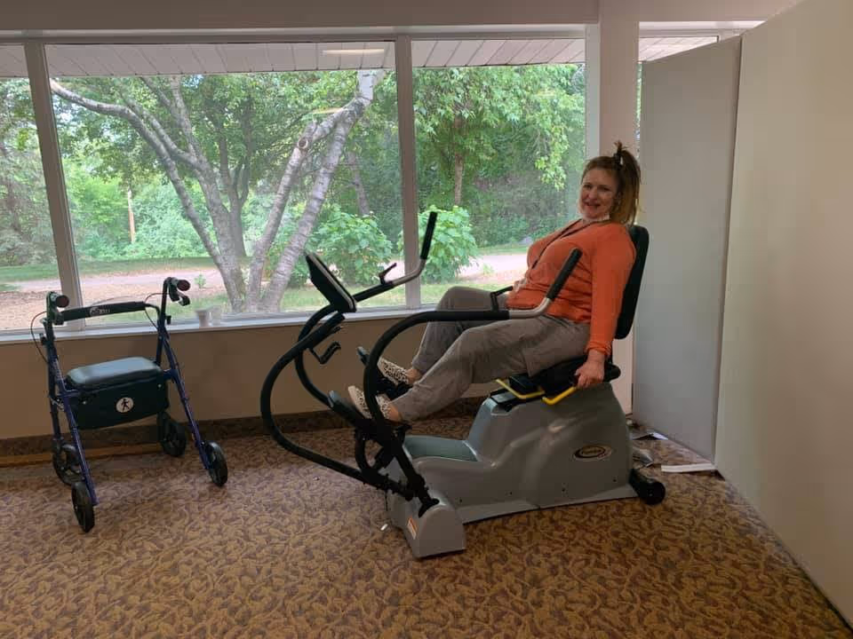 A woman in an orange top and gray pants is sitting on a recumbent exercise bike inside a room with large windows showing green trees outside. A blue walker is positioned nearby on a patterned carpet floor.