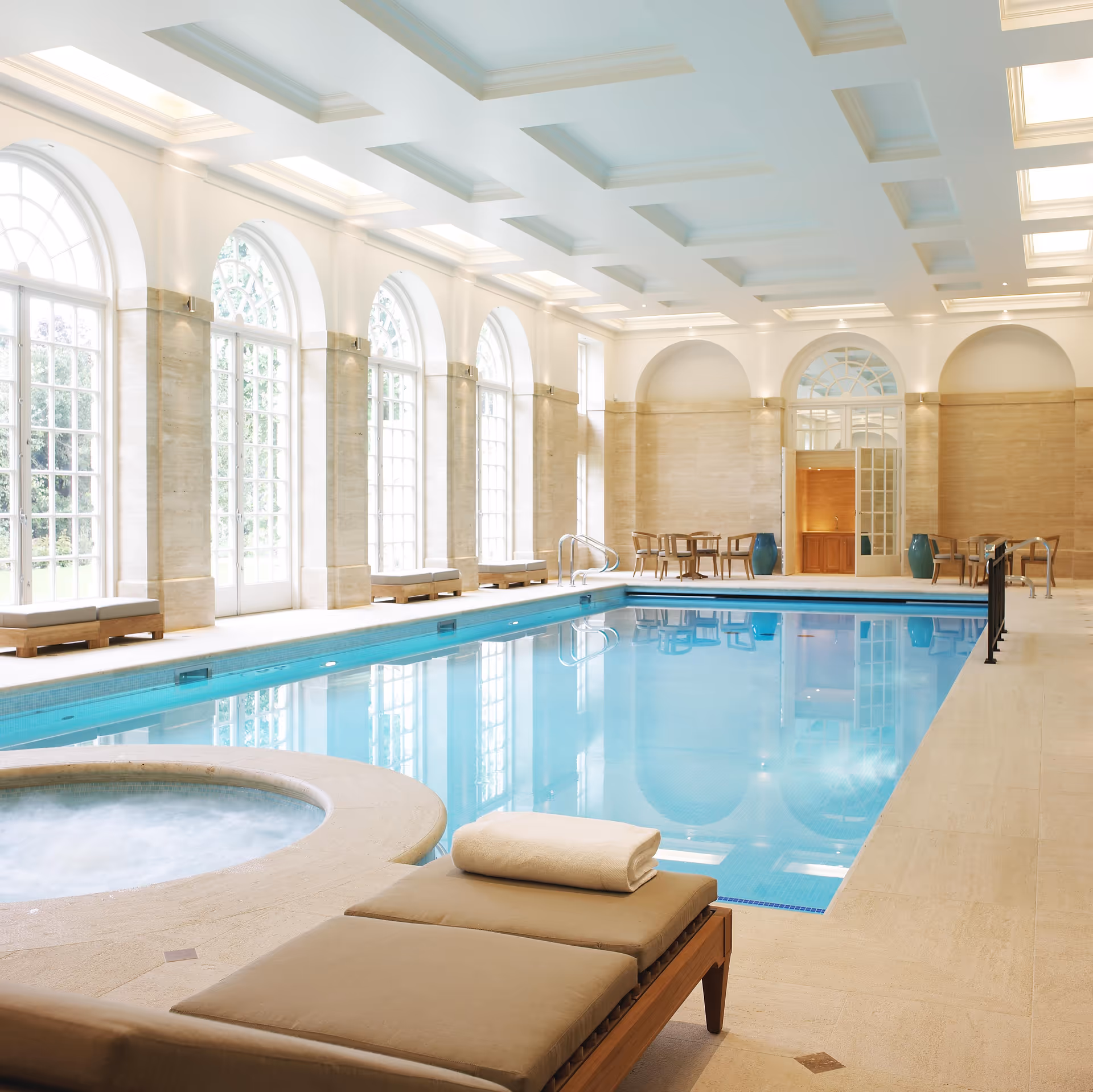 Indoor swimming pool area with large arched windows letting in natural light, beige tiled floors and walls, a hot tub in the foreground, cushioned lounge chairs with a rolled towel, and tables with chairs in the background.