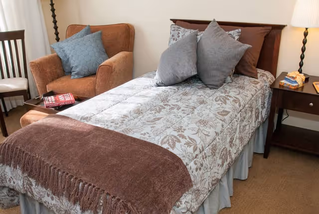 A cozy bedroom featuring a single bed with a floral patterned bedspread, three gray pillows, and a brown throw blanket. Next to the bed is a wooden nightstand with a lamp, a box of tissues, and a book. There is a brown upholstered armchair with a blue cushion and a wooden chair in the corner.