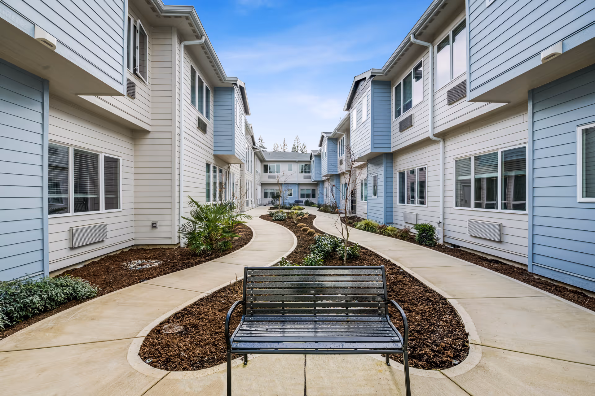 Outdoor courtyard area of a senior living facility with a black metal bench in the foreground, curved concrete walkways, landscaped garden beds with small plants and shrubs, and two-story buildings with blue and white siding on either side under a partly cloudy sky.