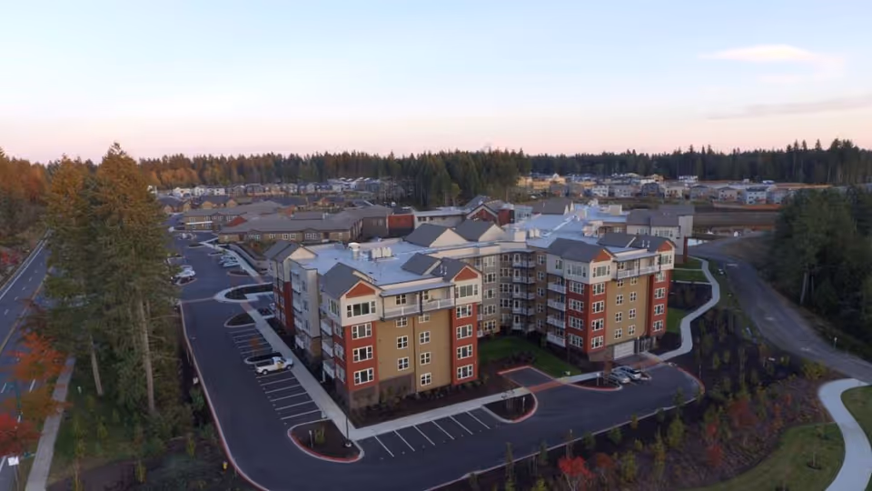 Aerial view of a multi-story senior living facility named Heron's Key surrounded by parking lots, trees, and landscaped pathways with a residential neighborhood and forested area in the background under a clear sky at dusk.