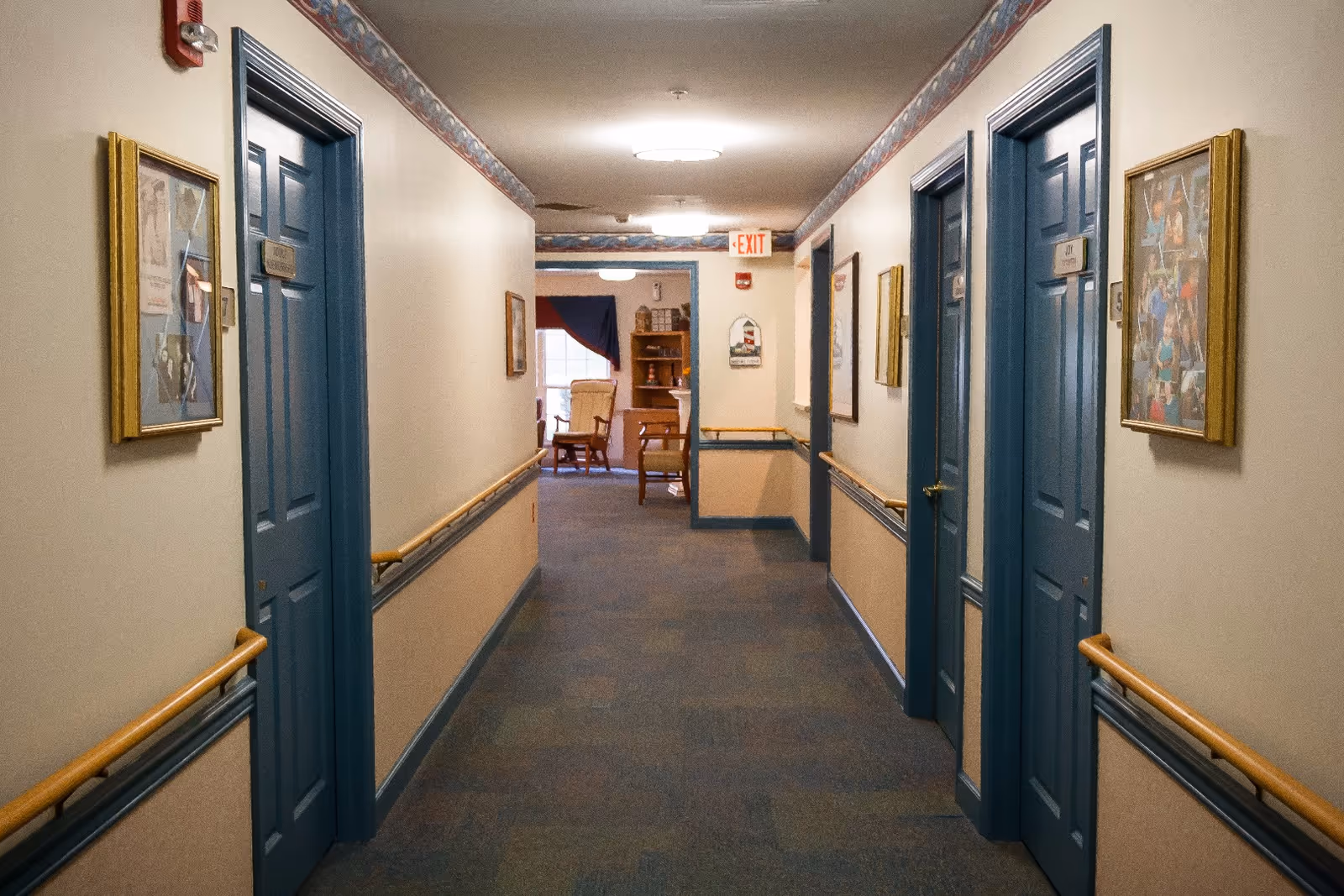 A carpeted hallway in a senior living facility with blue doors on both sides, beige walls with wooden handrails, framed pictures hanging on the walls, and a room visible at the end of the hallway with chairs and a window.