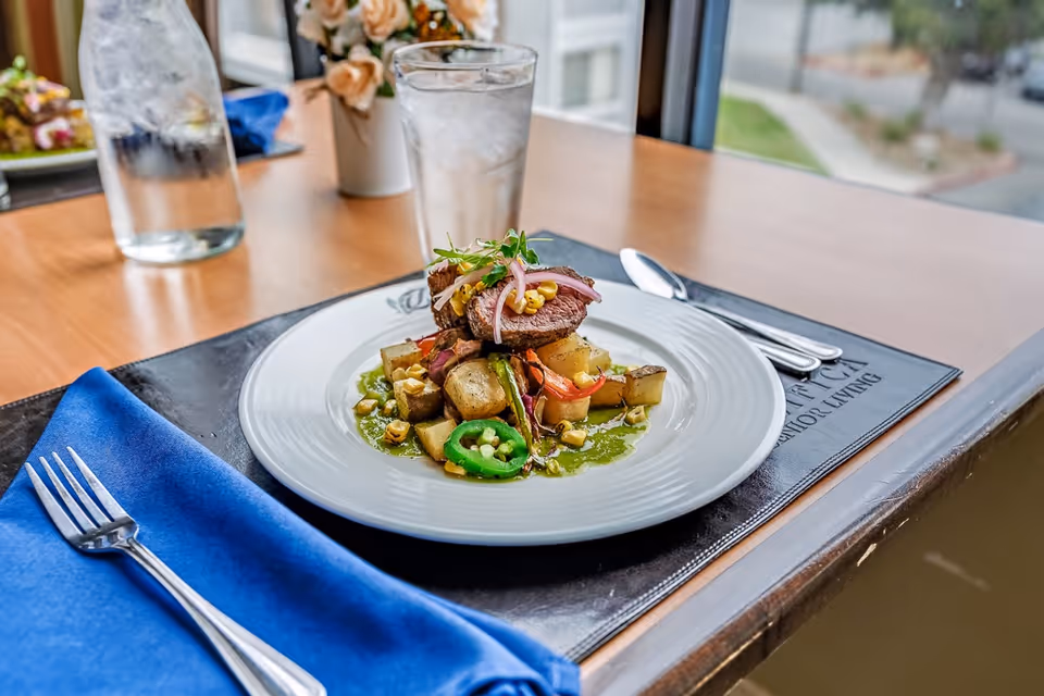 A plated gourmet meal consisting of sliced steak, roasted potatoes, jalapeño slices, corn, and garnishes on a white plate, set on a table with a blue napkin and silverware. A glass of ice water and a water bottle are also on the table near a window with a view outside.
