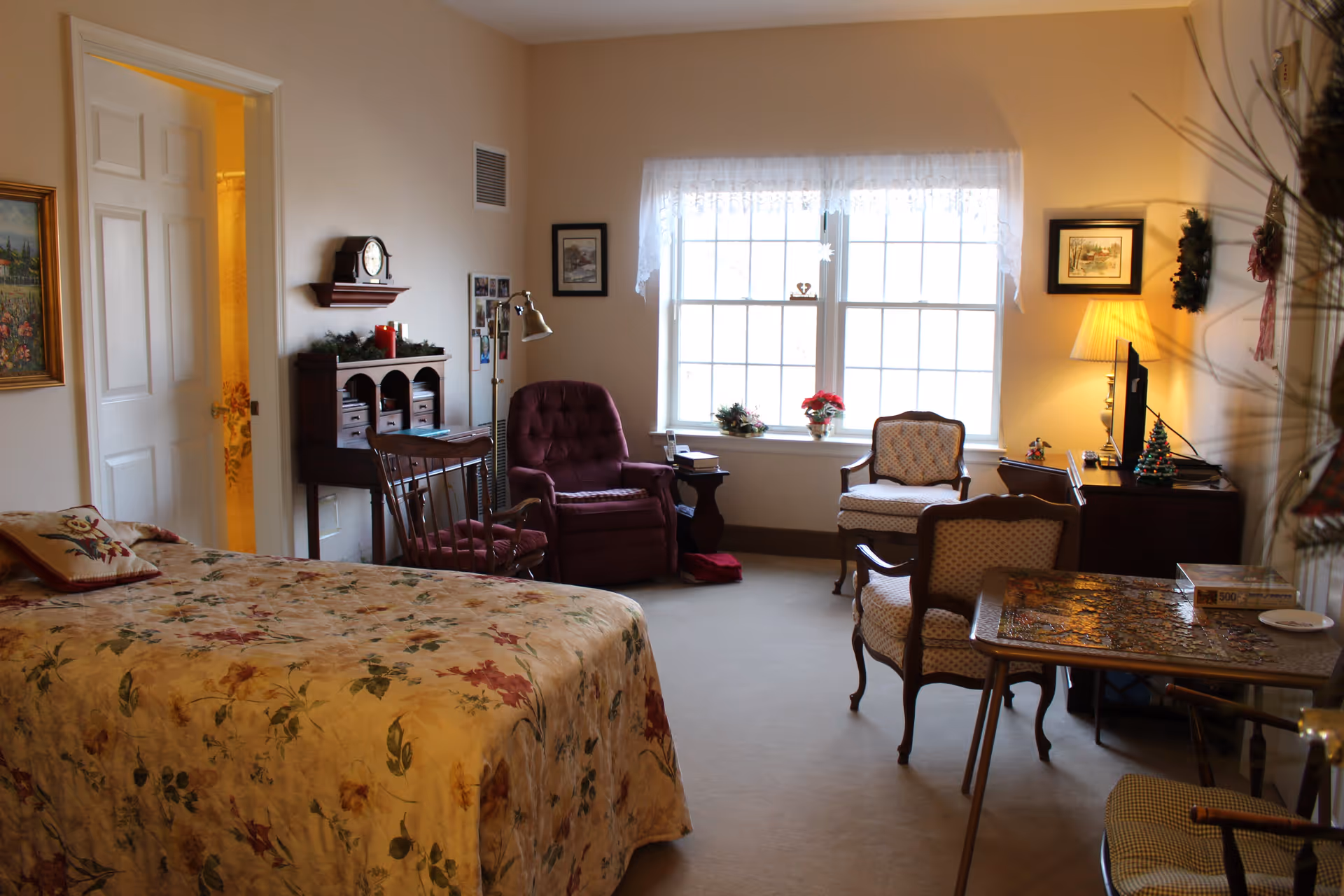 A cozy bedroom in a senior living facility with a floral bedspread on the bed, a wooden rocking chair, a burgundy recliner, two upholstered chairs, a small table with a puzzle in progress, a TV on a wooden dresser, and a window with lace curtains letting in natural light. The room is decorated with framed pictures, a clock on a wall shelf, and small holiday decorations.
