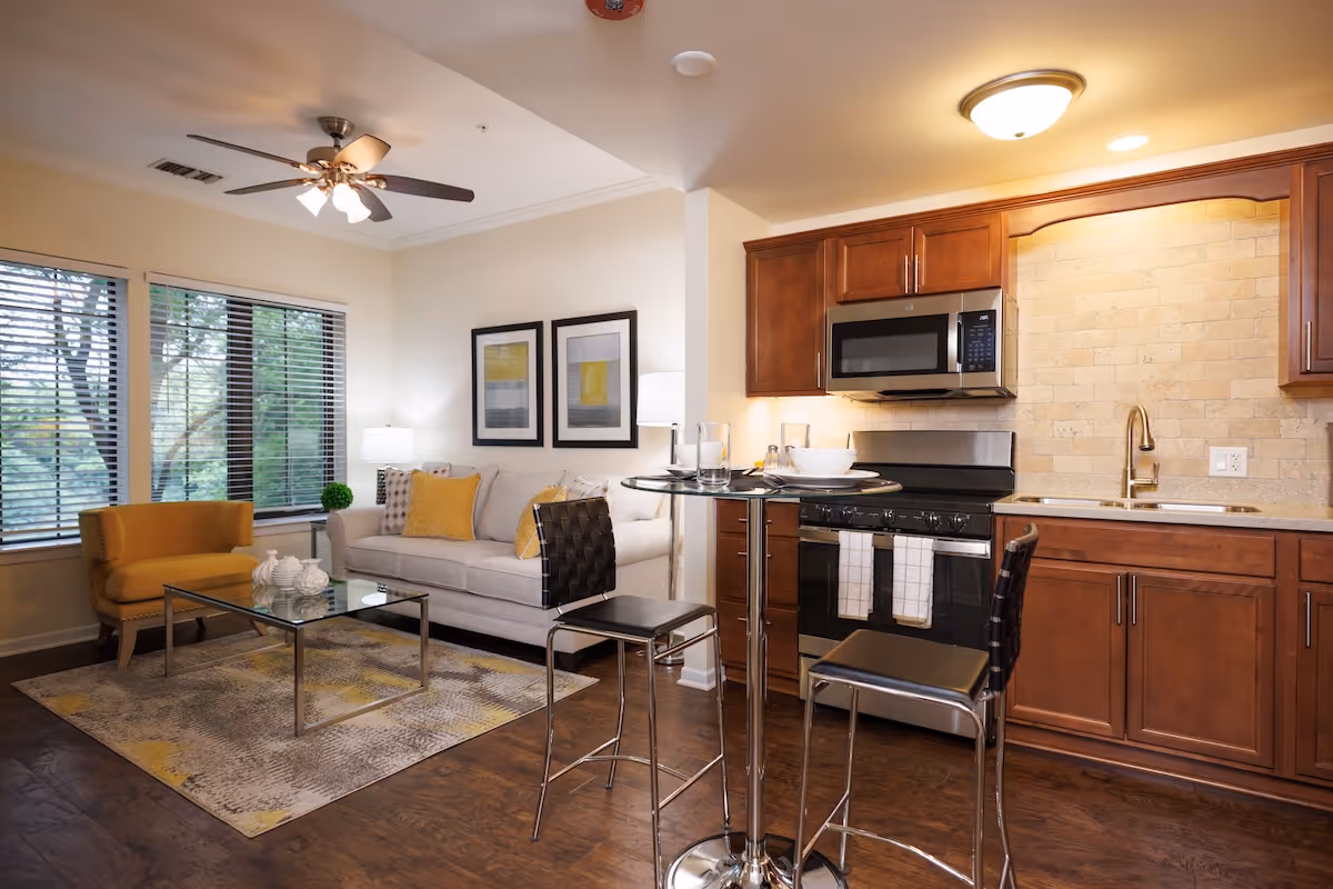 A cozy living area and kitchen in an apartment at Village At Victory Lakes. The living area features a beige sofa with yellow and patterned cushions, a yellow armchair, a glass coffee table on a patterned rug, and two framed artworks on the wall. The kitchen has wooden cabinets, a stainless steel microwave, stove, and sink, with a small round glass dining table and two black chairs in front.