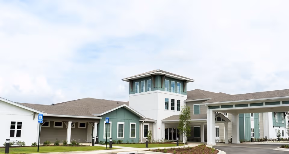 Exterior view of a senior living facility named The Canopy at Walden Woods, featuring a modern building with a mix of white and green siding, multiple windows, a covered entrance, and a tower-like structure in the center under a partly cloudy sky.