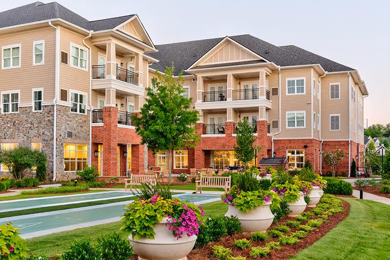 Exterior front view of a multi-story senior living building with balconies, landscaped gardens, large planters and benches.