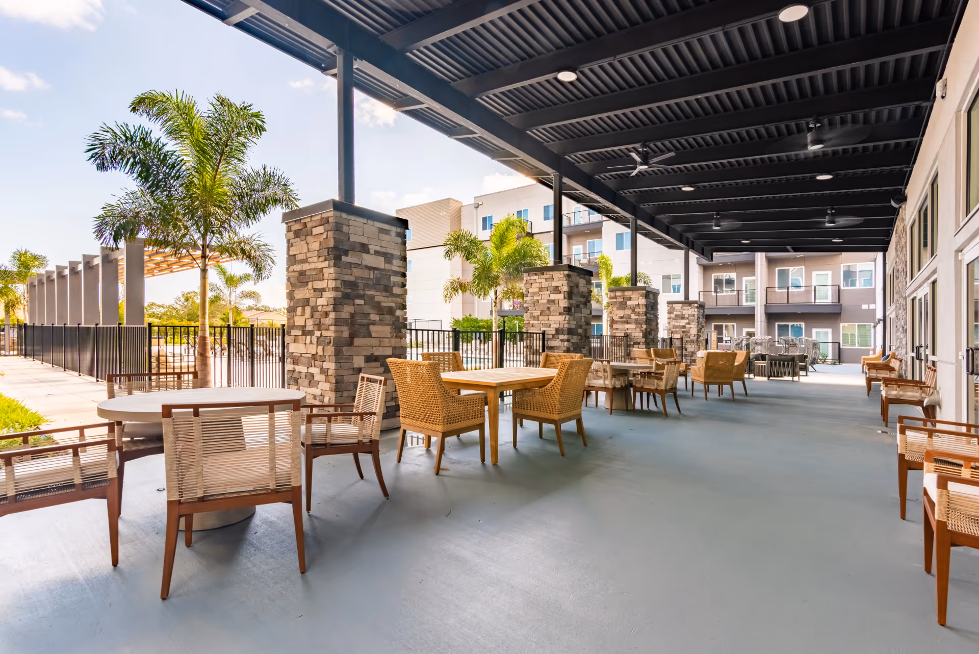 Covered outdoor patio with tables and chairs, stone pillars, and palm trees next to a multi-story building.