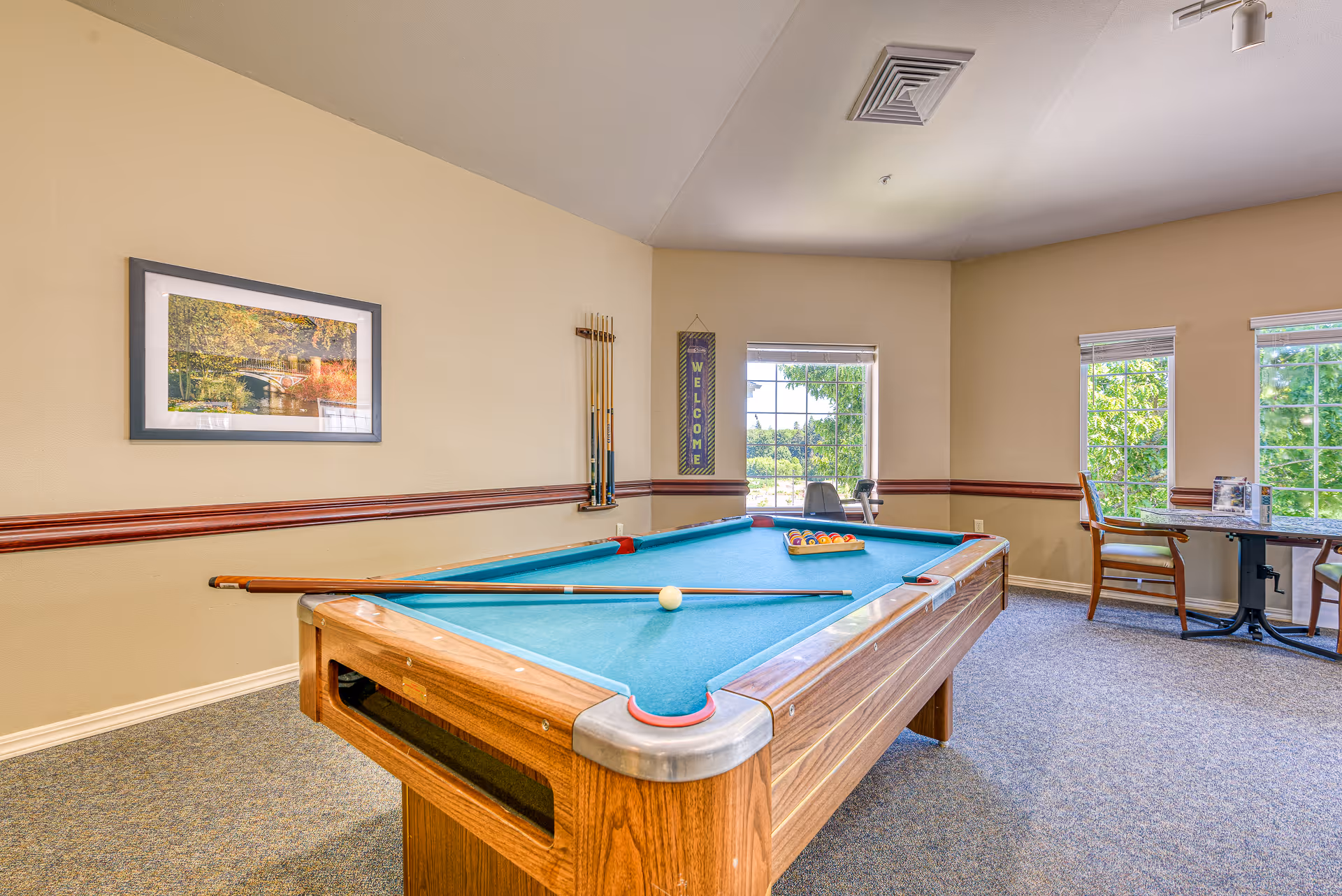 A recreational room with a pool table in the center, pool cues mounted on the wall, a framed picture, and a welcome sign. There are windows letting in natural light and a small table with chairs near the windows.