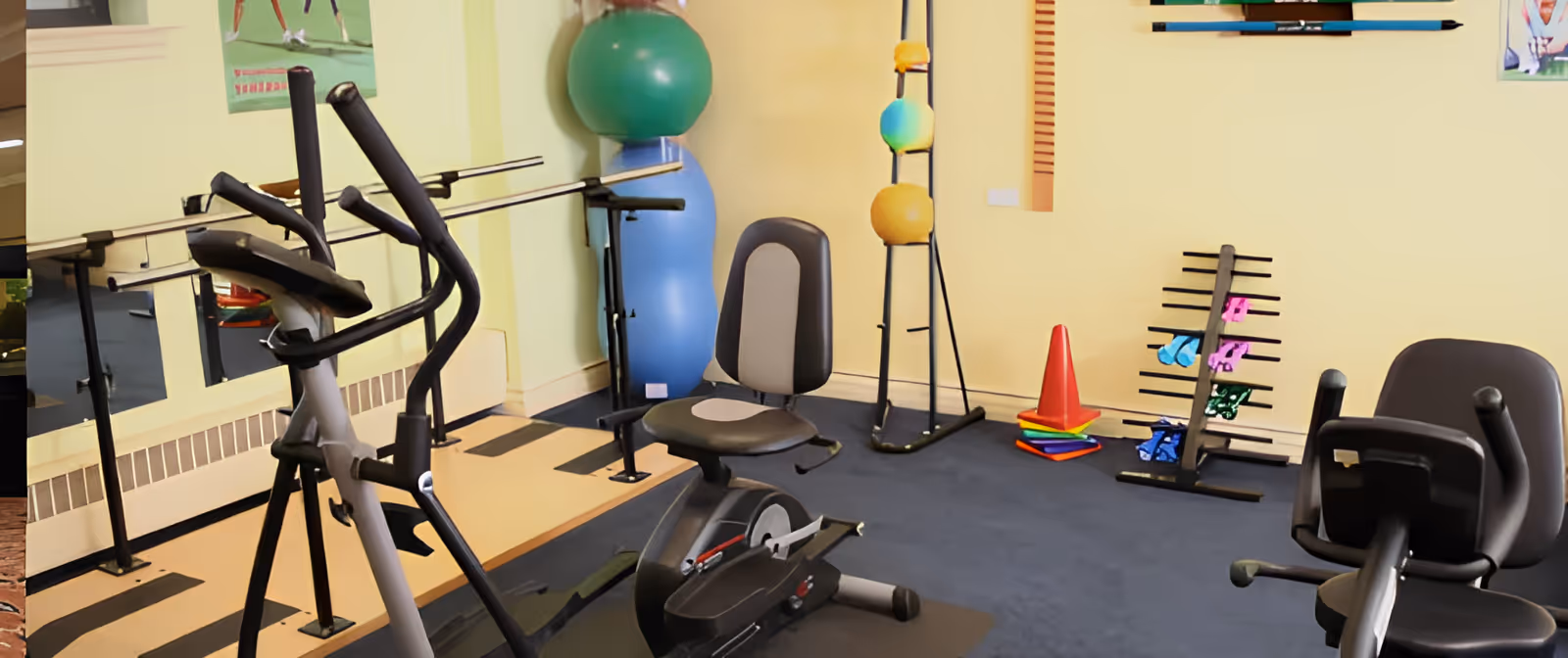 A small exercise room with various fitness equipment including stationary bikes, an elliptical machine, exercise balls, colorful cones, and weights. The room has a mirror on one wall and a light yellow paint color.