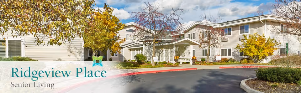 Exterior view of Ridgeview Place Senior Living facility showing a two-story building with beige siding, multiple windows, and a covered entrance. The surrounding area features autumn trees with yellow and orange leaves, landscaped bushes, and a paved driveway with a red curb.