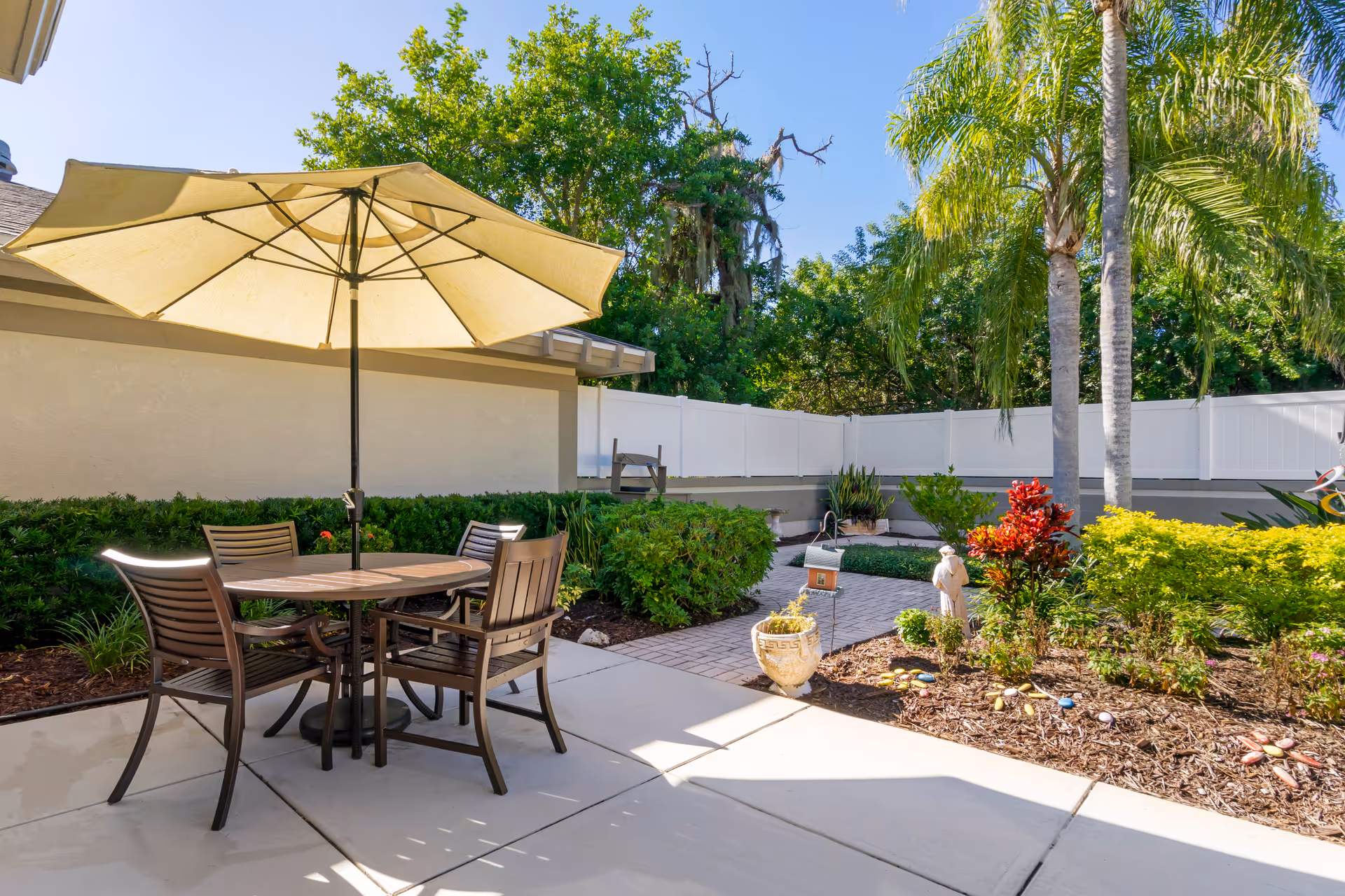 Sunlit patio with a round table, umbrella and chairs beside landscaped gardens, palm trees and a white fence.