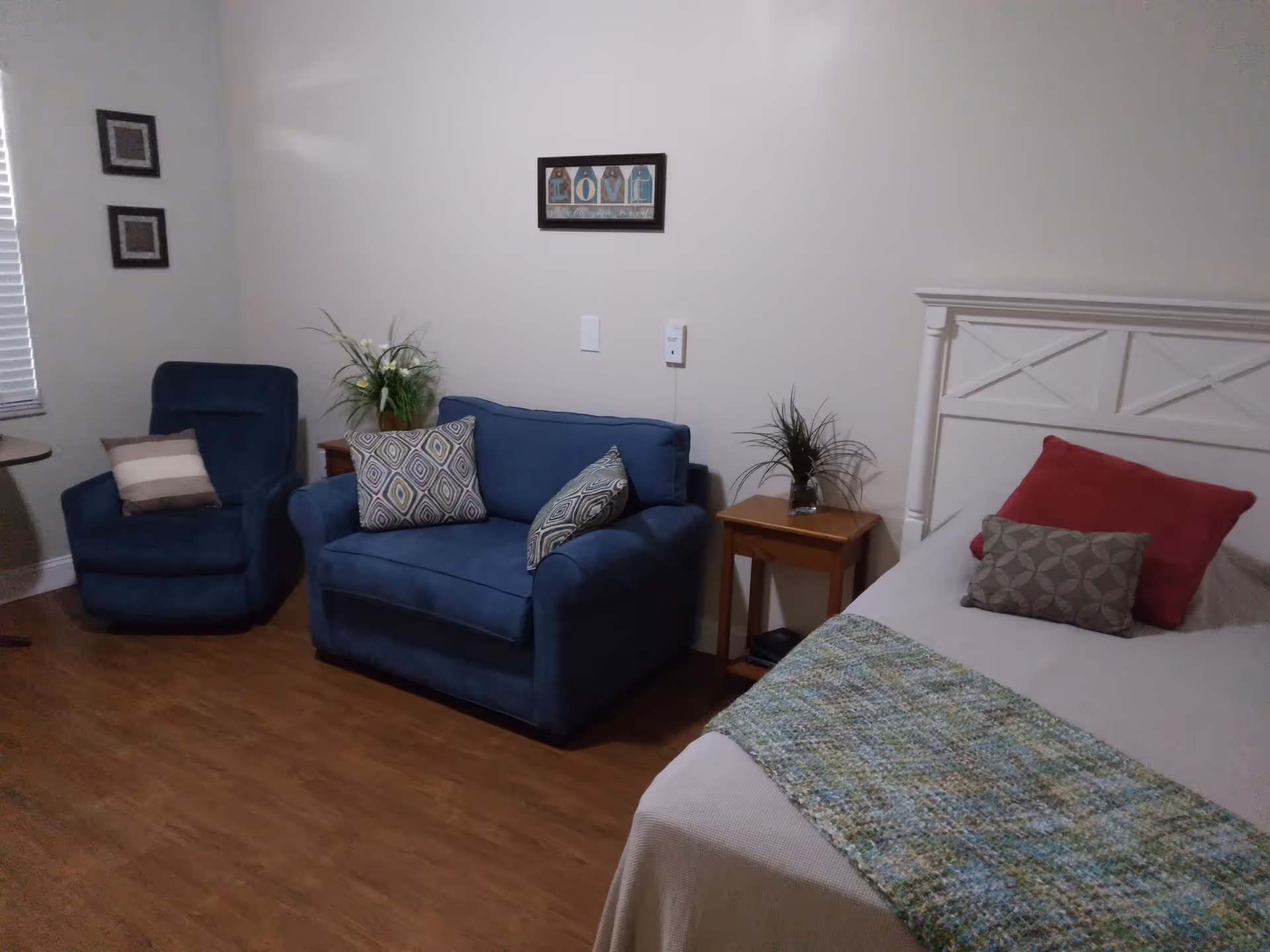 A cozy bedroom with a white bed featuring red and patterned pillows and a greenish-blue throw blanket. Next to the bed is a wooden side table with a small plant. Across from the bed is a blue loveseat with patterned cushions and a matching blue armchair with a striped pillow. The room has wooden flooring, light-colored walls, and framed artwork on the walls.