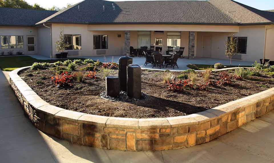 Outdoor courtyard area at Dublin Glenn Memory Care featuring a raised stone planter with various plants and a small water fountain in the center. Surrounding the planter is a concrete walkway, and in the background, there is a building with multiple windows and a patio area with several chairs and tables.