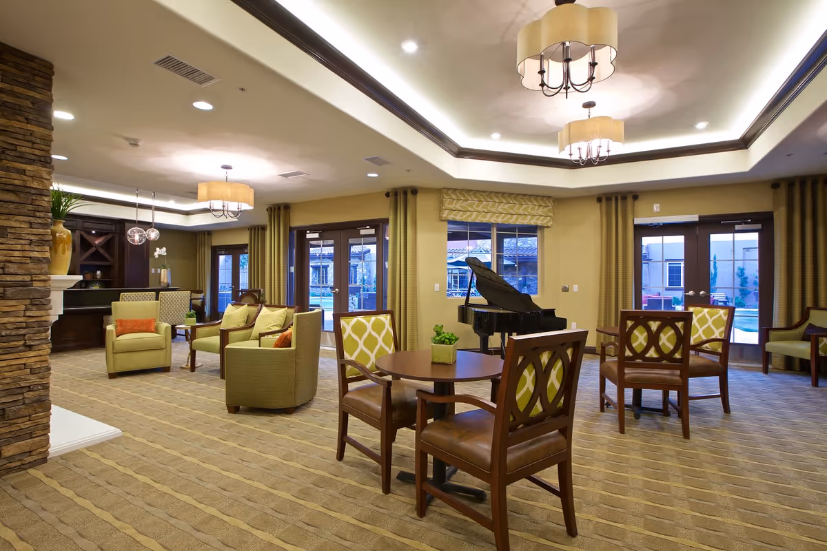 A spacious and well-lit common area with comfortable seating including armchairs and wooden chairs around small round tables. The room features a grand piano near a window, large windows and glass doors with curtains, and warm beige walls with recessed lighting and decorative ceiling fixtures. There is a stone pillar on the left side and a built-in dark wood shelving unit in the background.