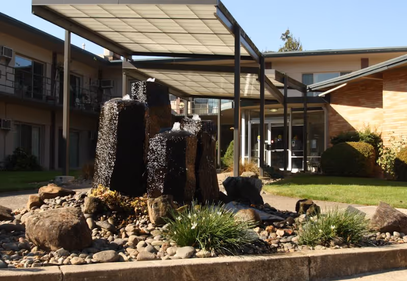Outdoor courtyard area of a retirement facility featuring a water fountain with multiple vertical stone pillars surrounded by rocks and plants, with a covered walkway and building entrances in the background.