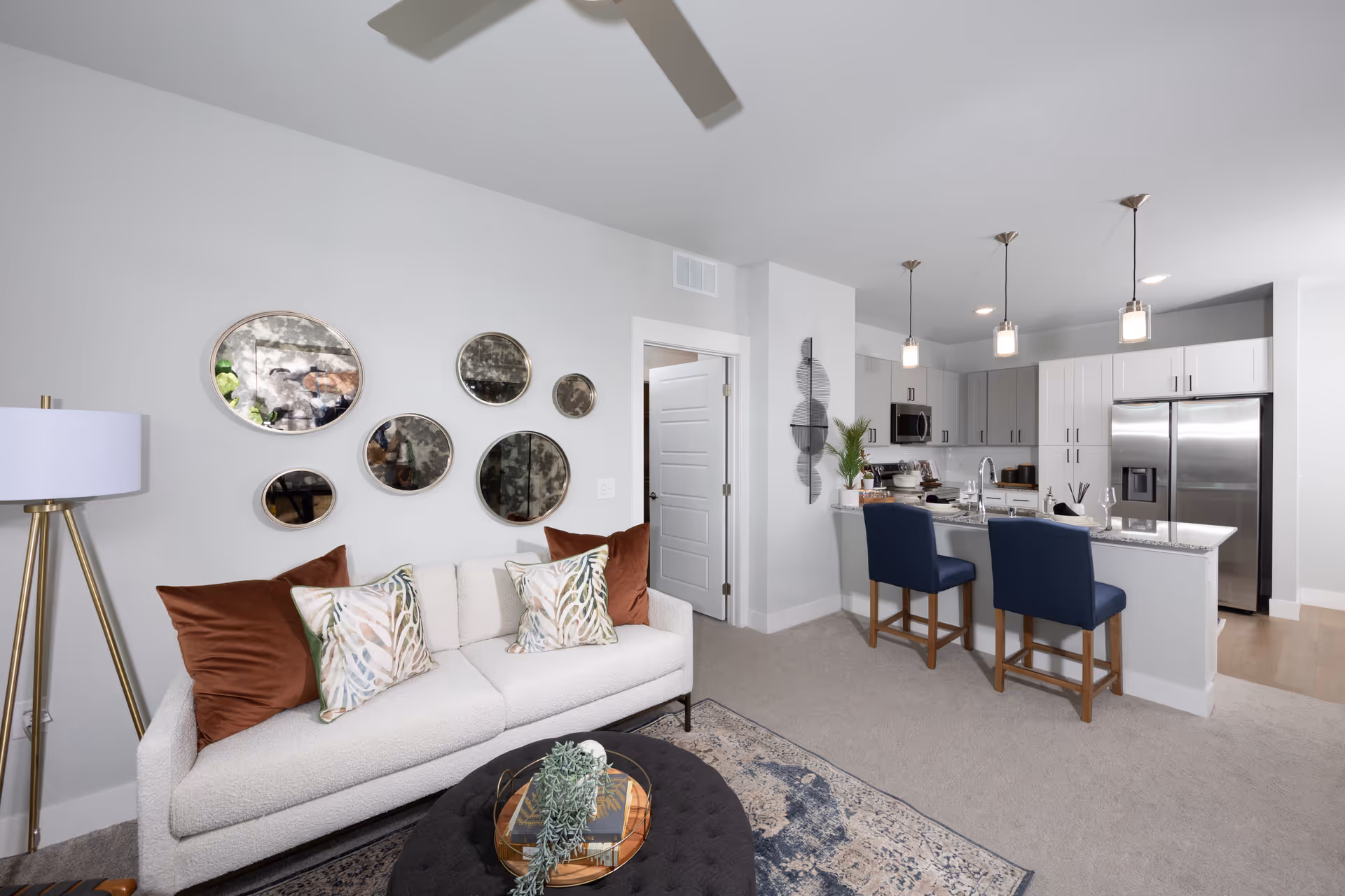 A modern living room and kitchen area with a white textured sofa adorned with brown and patterned pillows, a round black ottoman with a decorative tray and greenery, a floor lamp with a white shade, and a wall decorated with six round mirrors of varying sizes. The kitchen features white and gray cabinetry, stainless steel appliances including a refrigerator and microwave, a granite countertop island with a sink, and two blue upholstered bar stools. Three pendant lights hang above the kitchen island.