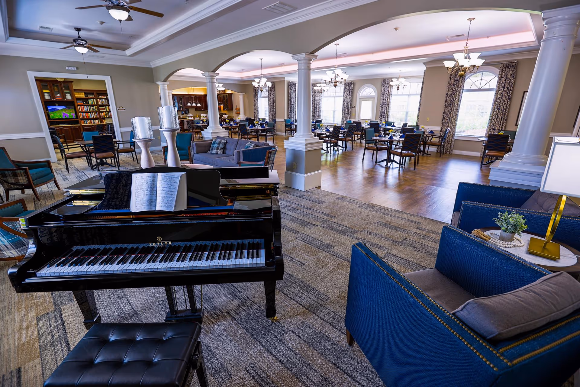 Interior view of a senior living facility common area featuring a black grand piano with sheet music, blue upholstered chairs, a small table with a lamp and plant, and a dining area with multiple tables and chairs under chandeliers. The space has large windows with patterned curtains and white columns separating the seating and dining areas.