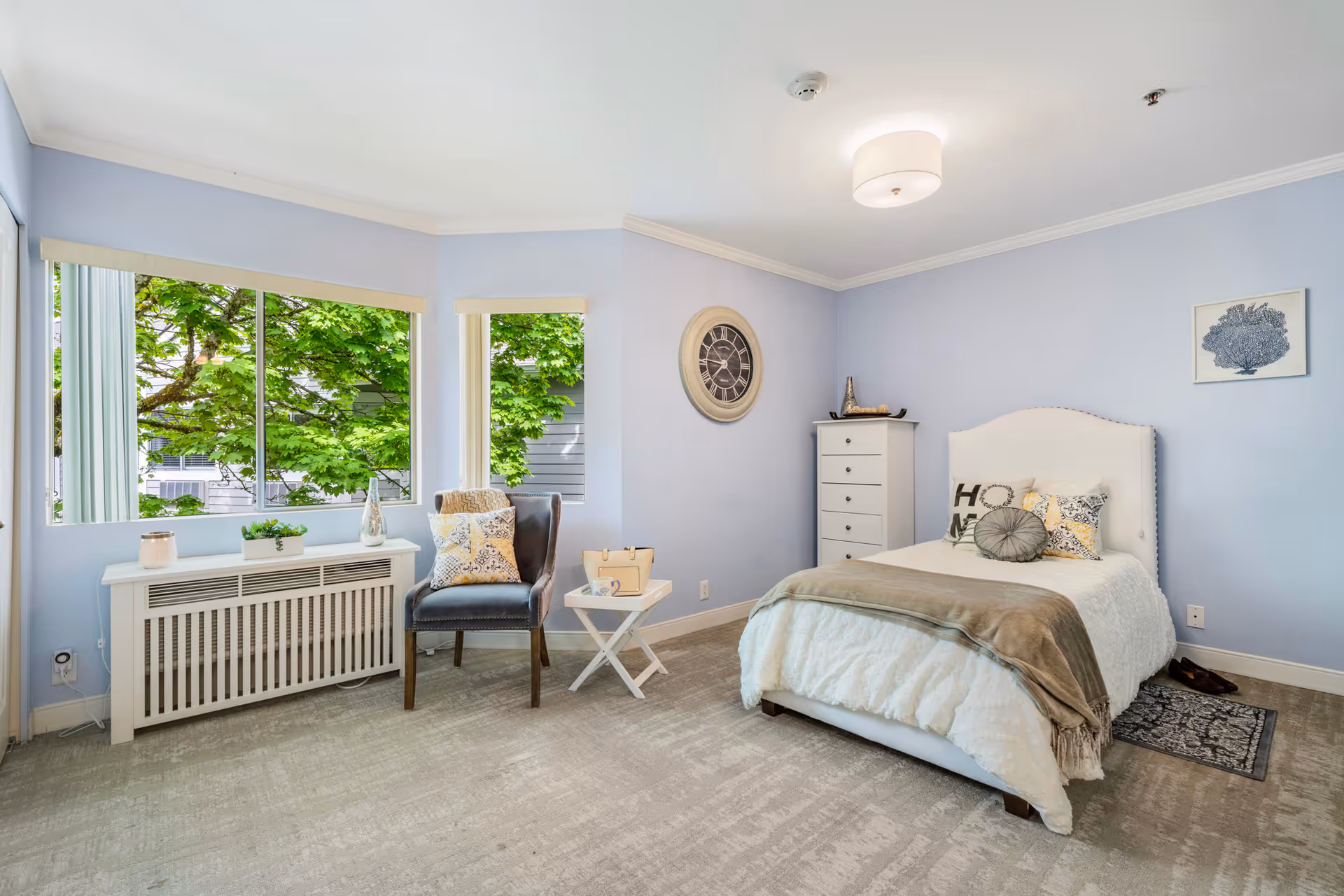 A bright and cozy bedroom with light blue walls, a large window showing green leafy trees outside, a white bed with multiple decorative pillows and a beige throw blanket, a white dresser, a small side table with a basket, a dark gray armchair with a patterned pillow, a wall clock, and a framed artwork on the wall.