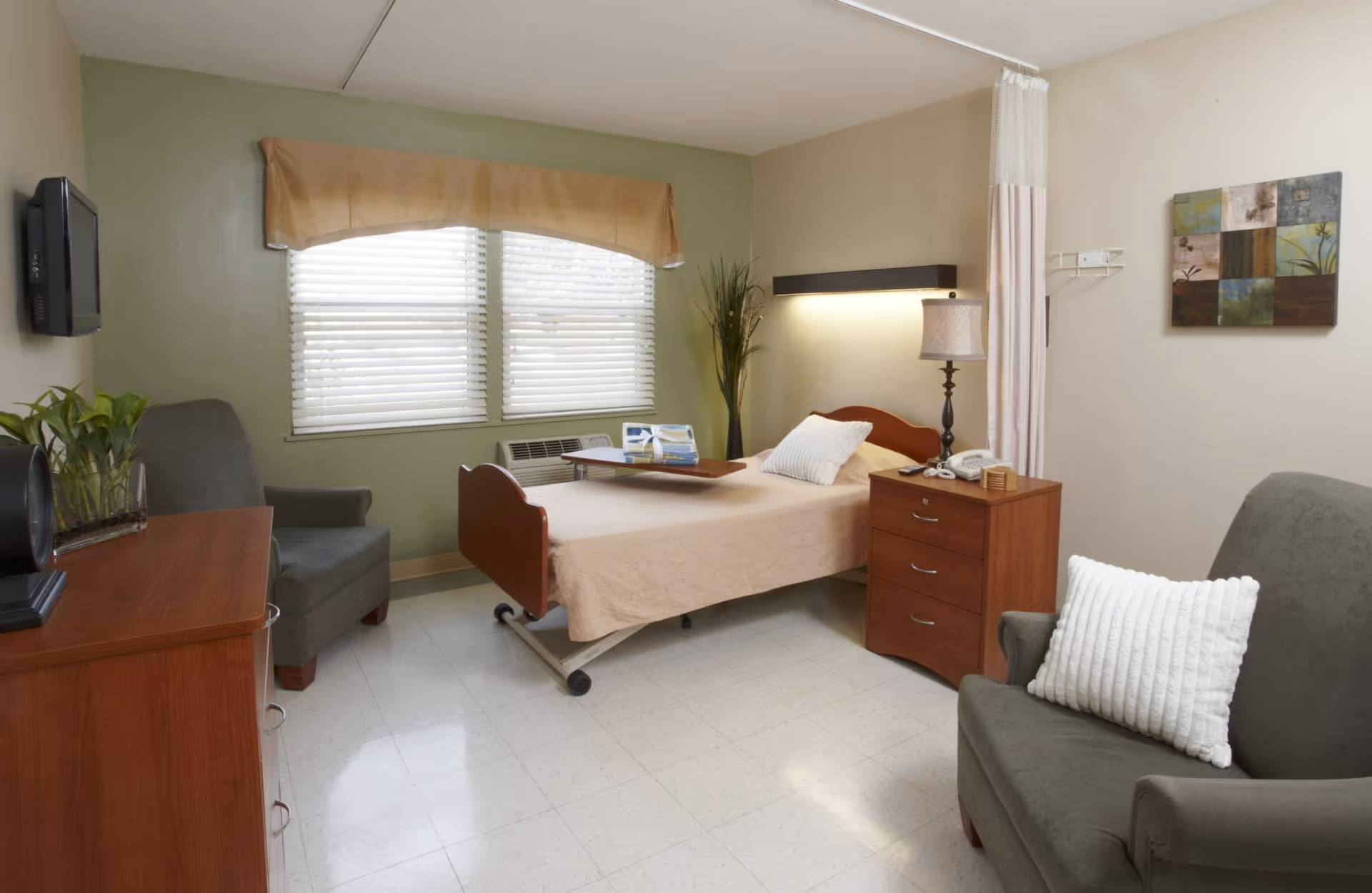 A bright single-occupancy senior care bedroom with a medical bed, nightstand, armchairs, wall-mounted TV, and a window with blinds.