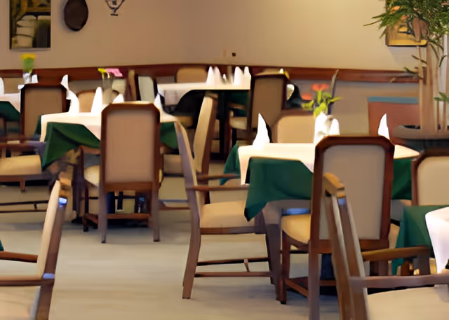 Interior view of a dining room with multiple tables covered in white and green tablecloths, each set with folded white napkins and small flower vases. Wooden chairs with cushioned seats surround the tables, and there is a large potted plant in the corner.
