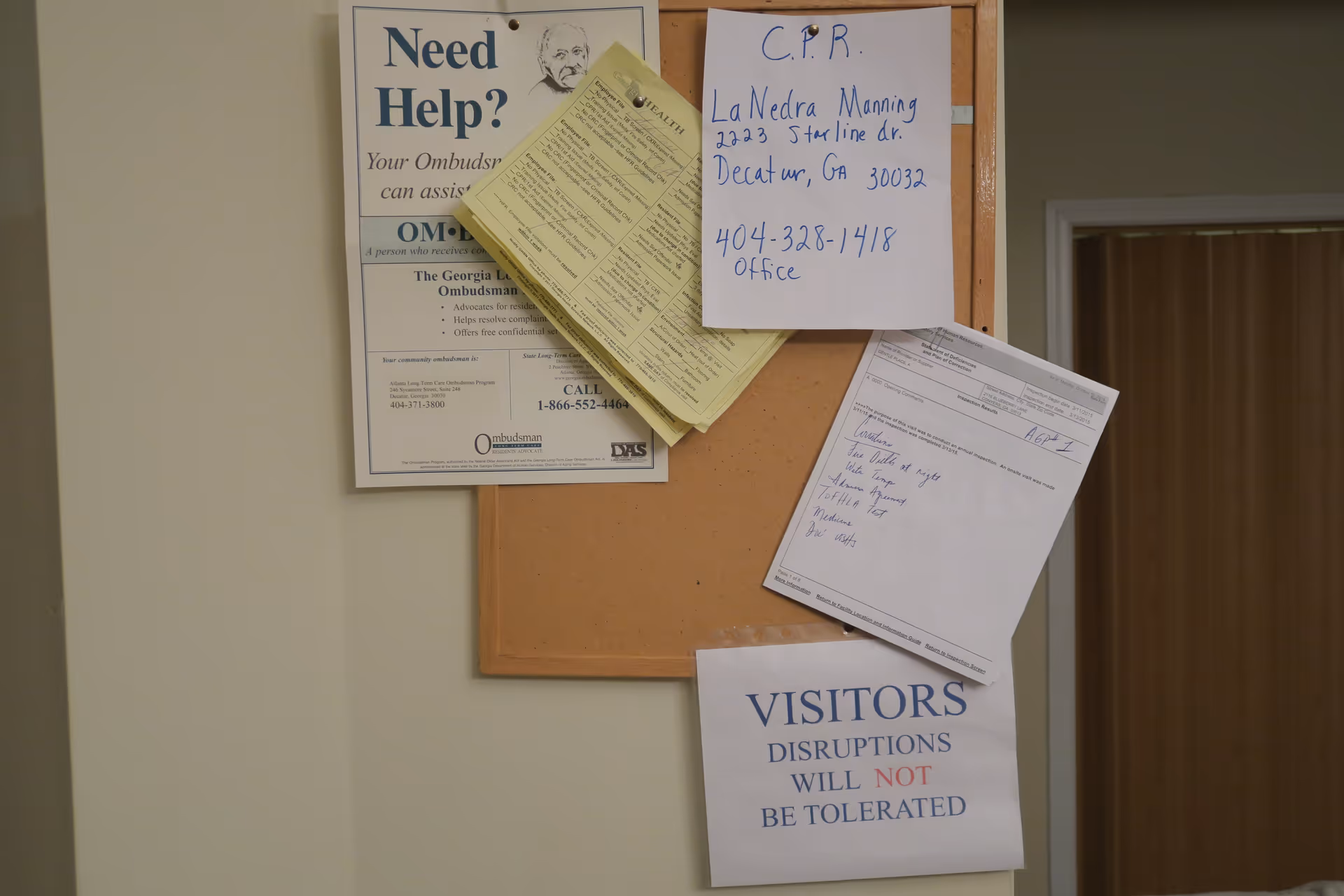 A bulletin board with multiple notices pinned on it, including a 'Need Help?' ombudsman information sheet, a handwritten note with CPR contact details for LaNedra Manning, a printed inspection results sheet, and a sign stating 'VISITORS DISRUPTIONS WILL NOT BE TOLERATED'. The bulletin board is mounted on a beige wall next to a doorway with a wooden door.