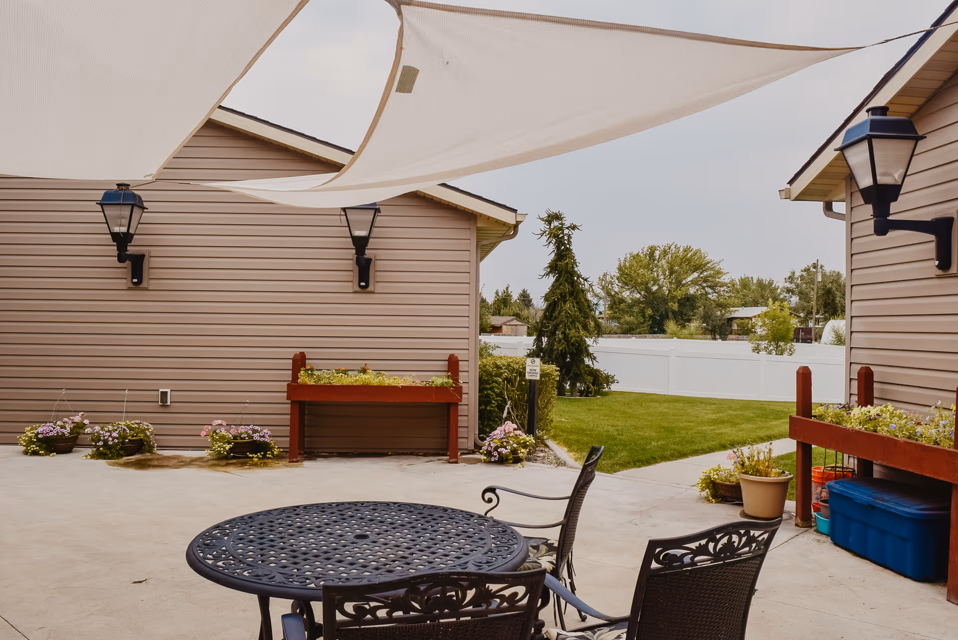 Outdoor patio area with a round metal table and chairs under white shade sails. The patio is surrounded by beige siding walls with mounted black lantern-style lights. There are flower pots and raised garden beds with plants, and a grassy area with trees and a white fence in the background.
