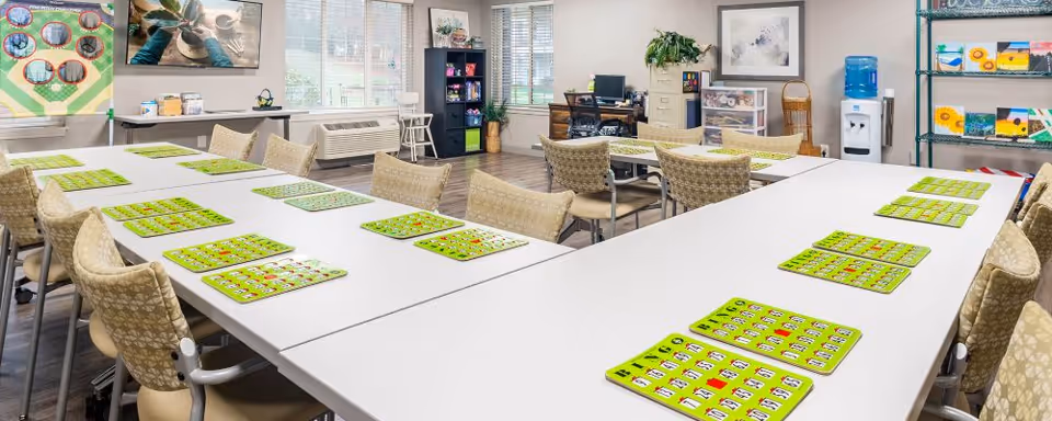 A bright and spacious activity room with several tables arranged in a U-shape, each table set with multiple bingo cards. Comfortable chairs surround the tables. The room has large windows letting in natural light, a water cooler, shelves with colorful paintings, and a bulletin board on the wall.