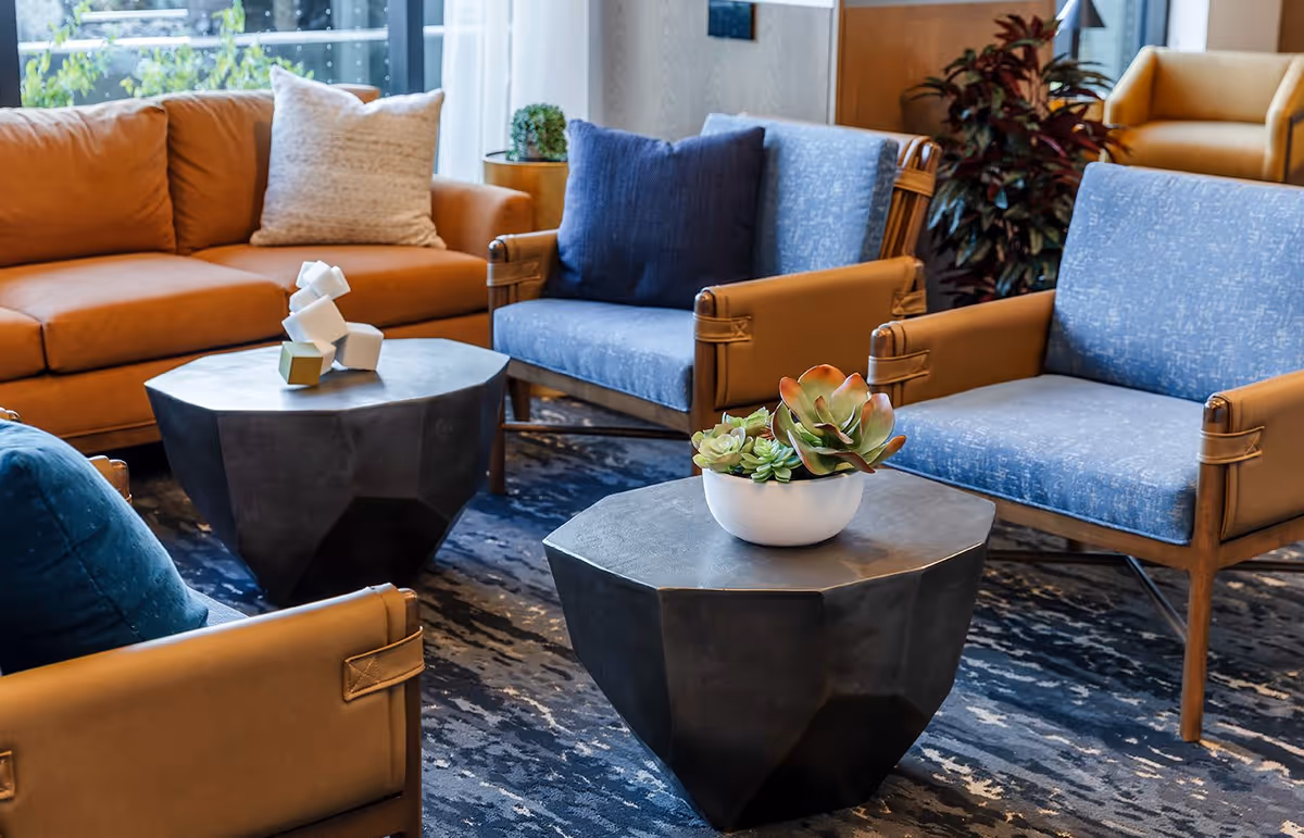 A cozy living room area with a brown leather sofa, two blue cushioned armchairs, and a matching blue loveseat. Two black geometric coffee tables are placed in the center, one with a white decorative sculpture and the other with a white bowl containing green and red succulents. The room has a blue patterned rug and a large window with sheer white curtains letting in natural light.