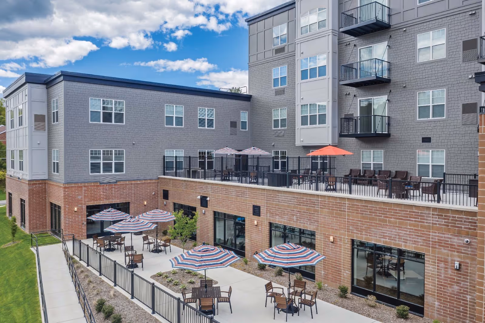 Outdoor patio area of Ridgecrest of McMurray facility with multiple tables and chairs under striped umbrellas on a concrete surface, surrounded by a black metal fence. The building exterior features a combination of brick and gray siding with multiple windows and balconies. The sky is partly cloudy.