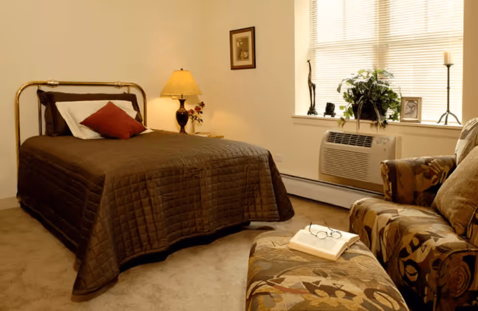 A cozy bedroom with a brass bed frame, brown quilted bedspread, and pillows. There is a bedside table with a lamp and flowers, a framed picture on the wall, and a window with blinds. In front of the window is an air conditioning unit. A patterned armchair and matching ottoman with an open book and glasses rest in the foreground.