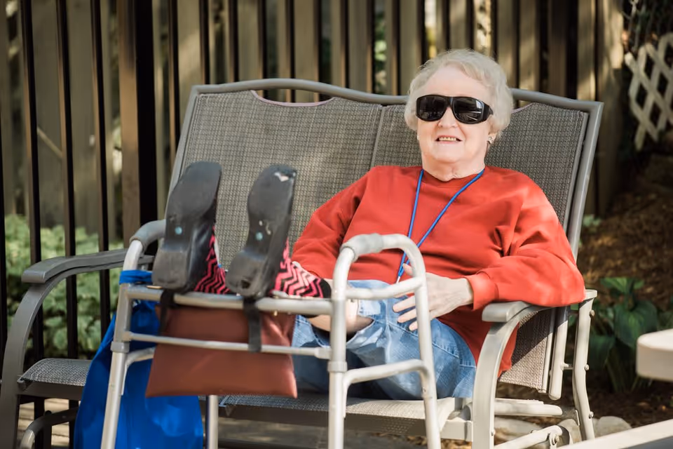 An elderly woman wearing sunglasses and a red sweater is sitting outdoors on a metal bench with her legs resting on a walker. She is smiling and appears relaxed in a garden or patio area with greenery and a wooden fence in the background.