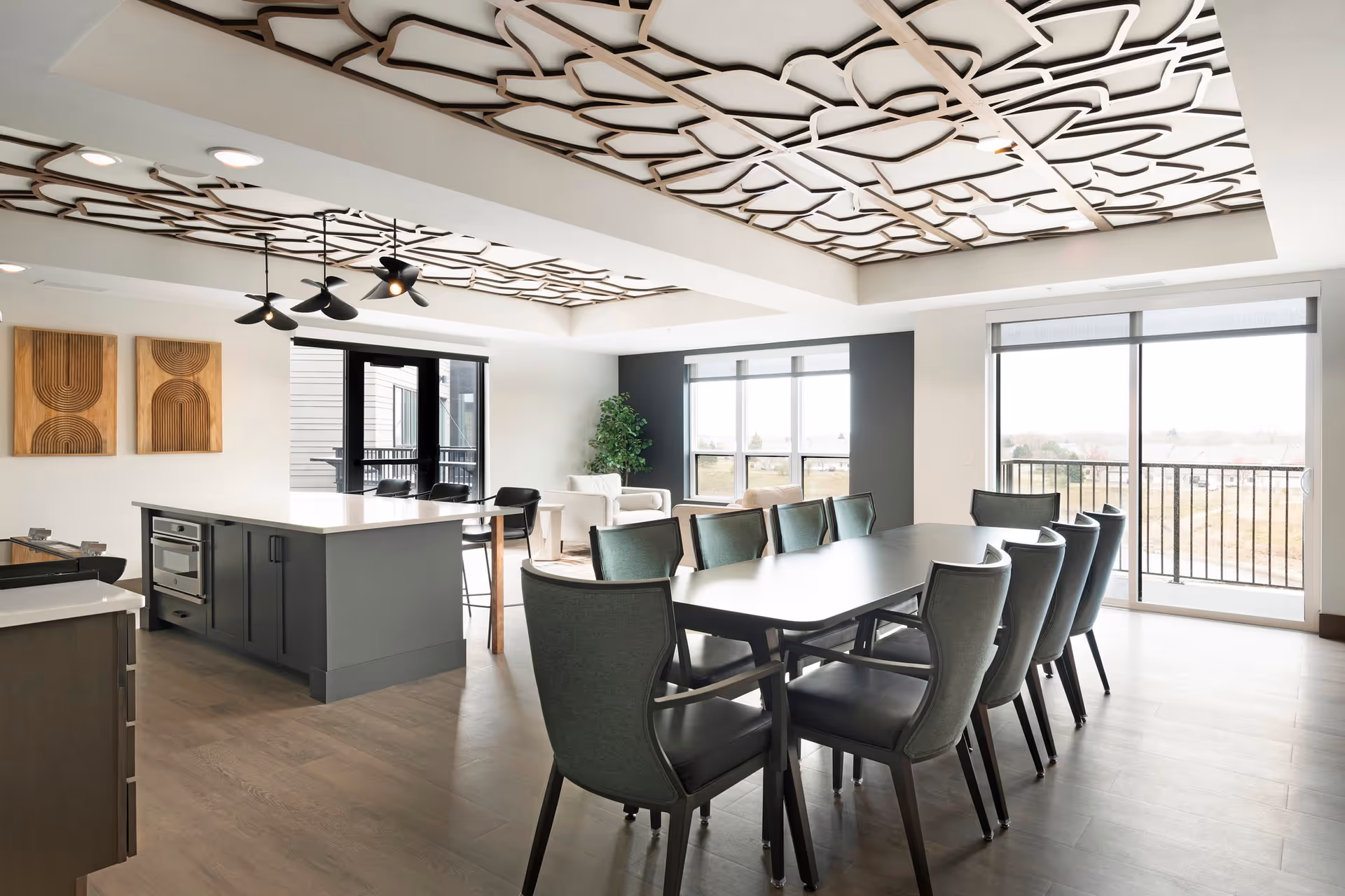 A modern, open-concept dining and living area with a long dining table surrounded by eight chairs, a kitchen island with bar stools, two armchairs near large windows, and decorative ceiling panels. The space is bright with natural light coming through large windows and sliding glass doors leading to a balcony.
