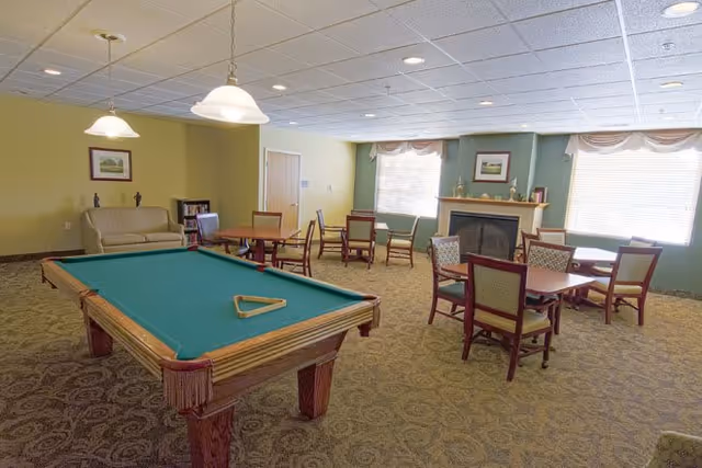 A spacious common room with a pool table in the foreground, several tables and chairs arranged around the room, a beige couch against the wall, two large windows with blinds, and a fireplace with decorative items on the mantel.