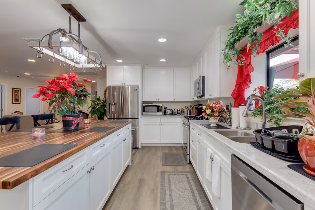 Bright and modern kitchen with white cabinets, stainless steel appliances including a refrigerator, microwave, stove, and dishwasher. A large wooden island with black placemats and a red poinsettia plant sits in the center. The kitchen has light wood flooring, recessed lighting, and a window with red curtains and green plants on the windowsill.