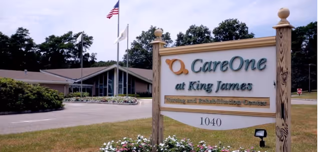 Exterior view of a senior living facility with a sign in the foreground that reads 'CareOne at King James Nursing and Rehabilitation Center 1040'. The building is single-story with a sloped roof, surrounded by trees and a driveway. Three flagpoles with flags are visible near the entrance.